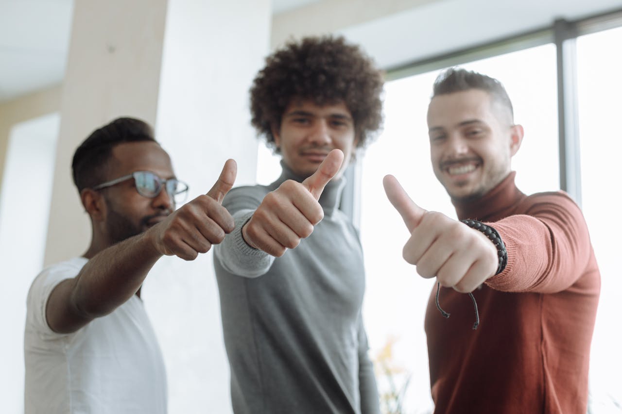 Three diverse men in a team give a thumbs up gesture indoors, symbolizing success and teamwork.